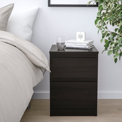Unha mesa de noite negra con caixóns, un vaso, libros e reloxo. Deseño minimalista xunto a unha cama con plantas e roupa neutra.