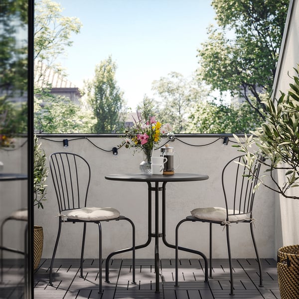 Conxunto de balcón moderno con mesa negra, coxíns brancos, flores coloridas e luces de corda.