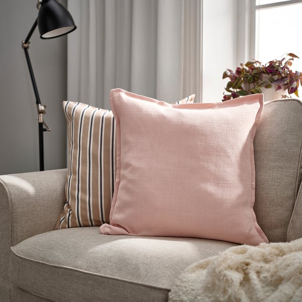 Sala de estar acolledora con sofá gris, dúas almofadas LAGERPOPPEL rosa clara, unha a raias, outra sólida, preto dunha lámpada e fiestra.