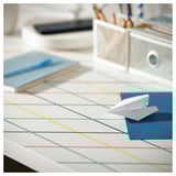Un escritorio branco cun patrín de cuadrícula colorido contén un avión de papel e un libro azul.