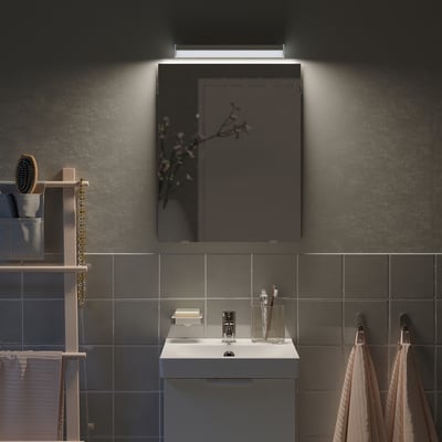 Pequeno espello gris de LÄRBRO no baño, iluminado sobre lavabo branco, rodeado de baldosas escuras.