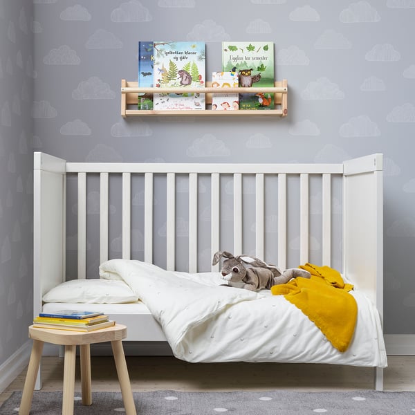 Habitación infantil con berce branco, peluche, manta amarela, taburete de madeira, libros, estante de parede con libros de cores e papel de papel de nube.
