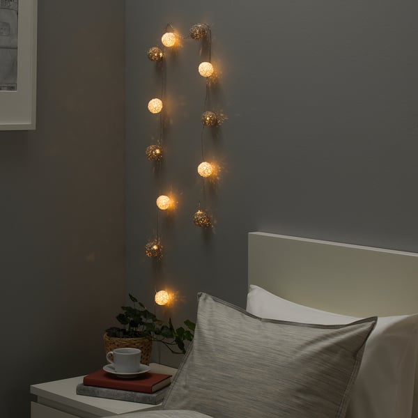Luces douradas LIVSÅR na parede gris. Cama quente con libro vermello, cunca e planta.