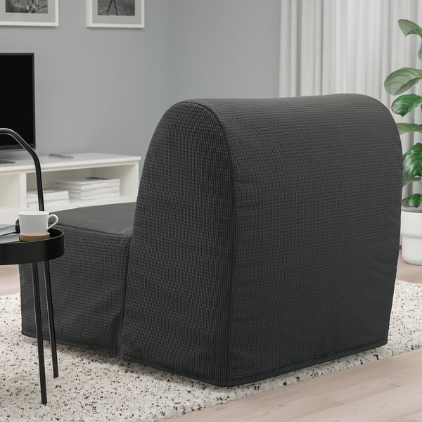 Cadeira-cama gris con deseño curvo, mesa lateral negra e cunca de café nunha habitación acolledora con gran xanela.