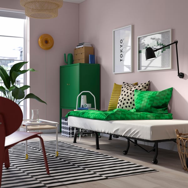 Dormitorio luminoso con paredes púrpuras claras, cama verde con almofadas brancas, alfombra a raias, cadeira vermella e unidade de almacenamento verde con libros e plantas.