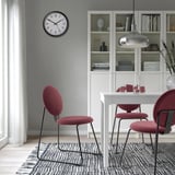 Zona de comedor moderna con 3 cadeiras tapizadas vermellas, mesa branca e alfombra gris. Gran reloxo de parede, armario branco con libros, elegante lámpada colgante.