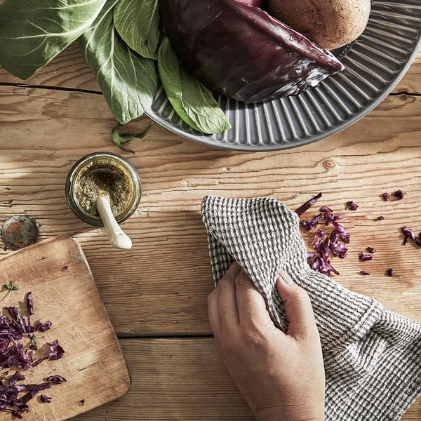 Unha man sostén unha toalla gris de prato de MARIATHERES, limpando unha mesa de madeira con repolo picado, verduras nunha bandexa e un frasco de especias preto.