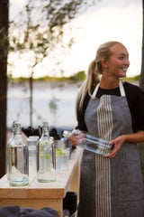 Unha muller cun mandil de raias brancas e negras serve bebidas ao aire libre. Ela sostén unha botella de vidro transparente e sorrí.