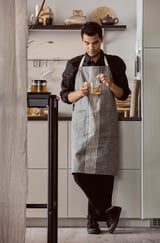Un home con camisa negra e mandil gris sostén unha cunca na cociña moderna. O mandil ten raias e bolsas. Estantes con elementos detrás.