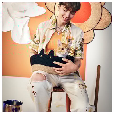 Persoa con gato en cesta de xute, camisa floral, jeans rasgados, sombreiros, paredes de cores.