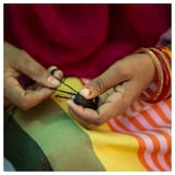 As mans con pulseiras vermellas tecen fíos negros nun téxtil de raias coloridas.