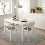 Mesa de comedor branca MELLTORP con 4 cadeiras de almofada negra, configuración de café, alfombra gris, nunha cociña luminosa con plantas.