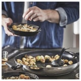 Escena da cociña: cociñar con tixola negra e espátula, verduras fritas na estufa, ferramentas e vaso de auga no mostrador.