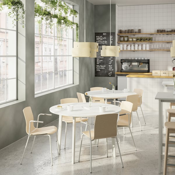 Café moderno con mesas redondas MITTZON, cadeiras de madeira clara, fiestras luminosas, plantas colgantes. Mesas pequenas, con capacidade para catro asentos cada unha.