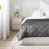 Dormitorio minimalista con paredes brancas, cama gris e pequena mesa de noite.