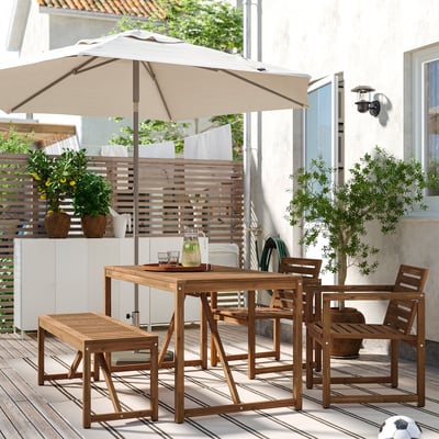 Patio exterior con mesa de madeira NÄMMARÖ, cadeiras, banco e paraugas beige. Rodeada de plantas e un edificio branco.