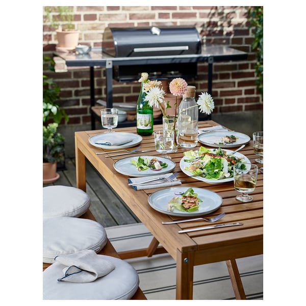Conxunto de comedor ao aire libre con mesa de madeira, cadeiras de almofada grises, pratos, vasos e flores.