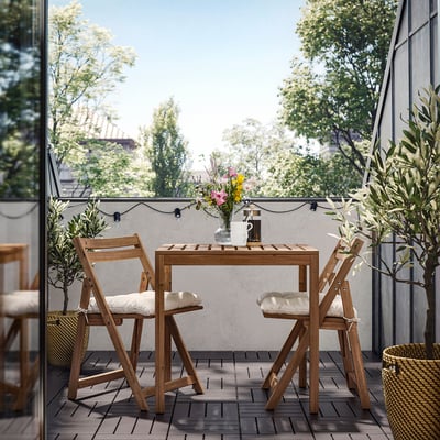 Patio con mesa NÄMMARÖ, cadeiras acolchadas brancas, plantas e luces.