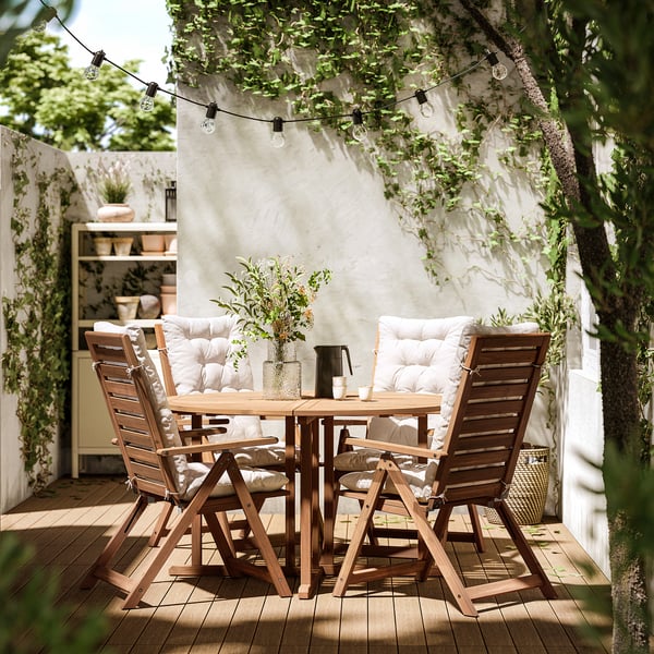 Patio exterior con mesa e cadeiras NÄMMARÖ, coxíns. Luces de corda colgan enriba da cabeza, plantas en pota, paredes brancas.