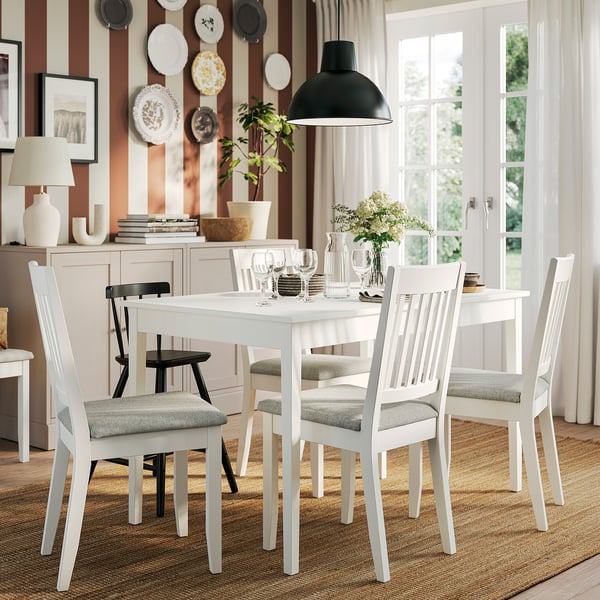 Mesa de comedor branca con 4 cadeiras grises, extensible, decorada con pratos, lámpada, libros e vasos.
