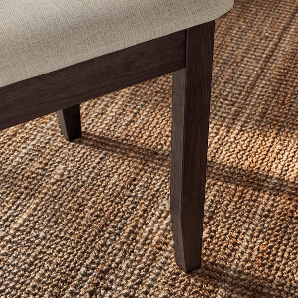 Primeiro plano da perna da cadeira NÄSINGE, madeira escura sobre alfombra texturizada. Robusto e elegante, mostrando detalles finos e construción de calidade.