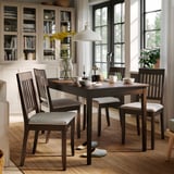 Zona de comedor acolledora con mesa NÄSINGE, cadeiras grises, flores, estante de libros e grandes fiestras.