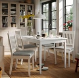 Comedor luminoso con mesa e cadeiras brancas NÄSINGE, coxíns grises, chan de madeira, gran xanela e estante.