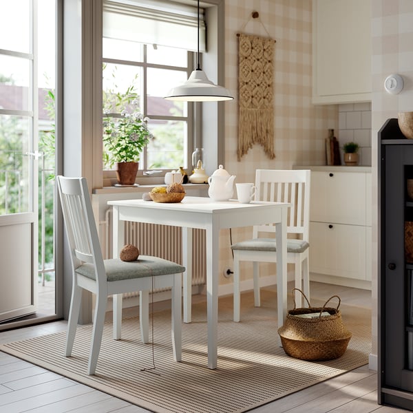 Recuncho de comedor acolledor con mesa branca, coxíns beige. Cestas e teteira sobre a mesa. Cociña con gran fiestra.