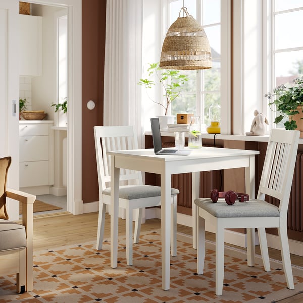 Espazo de comedor acolledor con mesa branca NÄSINGE, cadeiras de madeira e lámpada colgante de vimbio. Portátil, vidro e planta sobre a mesa. Compacto pero acolledor.