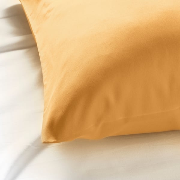 Funda de almofada amarela dourada sobre unha cama branca, tecido liso, peche de sobr. Textura suave, aspecto luxoso.