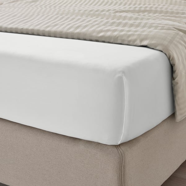 Unha cama ben feita con sabas brancas, cun tecido liso de seda deseñado para o confort. A saba axustada é visible baixo unha manta beige de raias.