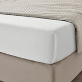 Unha cama ben feita con sabas brancas, cun tecido liso de seda deseñado para o confort. A saba axustada é visible baixo unha manta beige de raias.