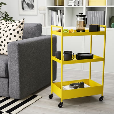 Carro rodante amarelo brillante NISSAFORS de 3 niveis na sala de estar xunto ao sofá gris. Contén tazas, pratos e cafeteira. Deseño compacto e versátil.