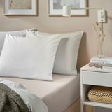 Un dormitorio cunha cama NONNEA en beige claro. A cama ten almofadas de algodón branco e unha manta. Unha mesa de noite cun vaso, libros e gafas está ao seu lado.