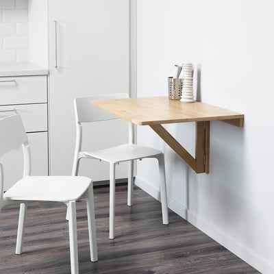 Mesa de cociña de madeira plegable con dúas cadeiras de plástico branco contra unha parede branca.