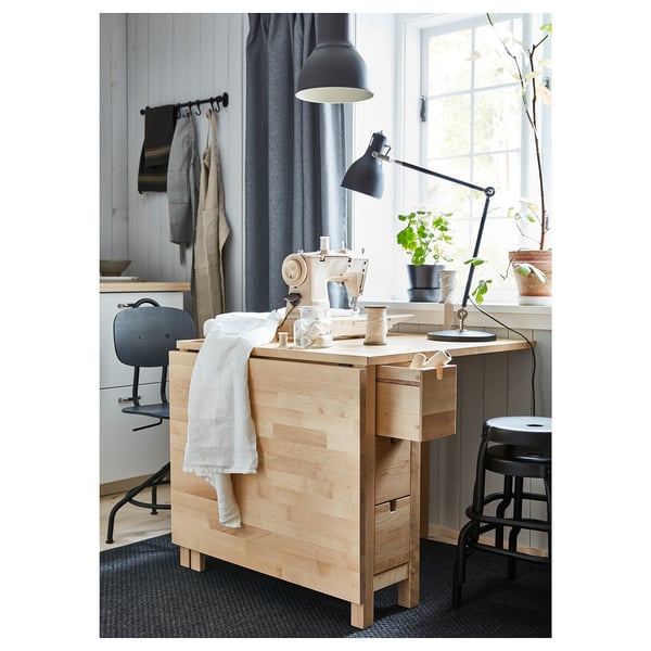 Sala de artesanía minimalista con escritorio de madeira clara, máquina de coser, taburete negro e lámpada axustable.