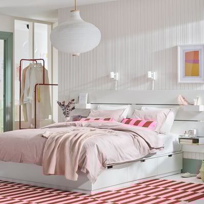 Dormitorio branco moderno con alfombra a raias, roupa de cama rosa, paralume colgante, estante de roupa e marco de cama branco NORDLI con caixóns de almacenamento.