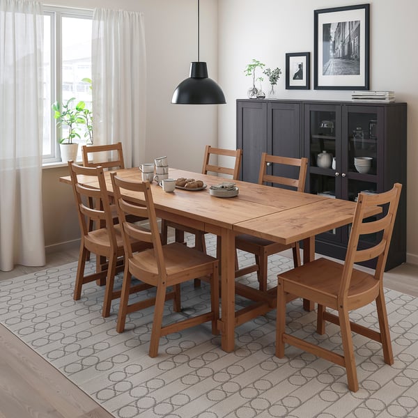 Comedor moderno con mesa NORDVIKEN, cadeiras e alfombra con patróns. Luz pendente negra, sideboard escuro e cortinas transparentes.