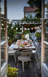 Acolledor conxunto de comedor ao aire libre con cadeiras brancas e mesa de madeira nunha cuberta, adornada con luces colgantes e plantas.