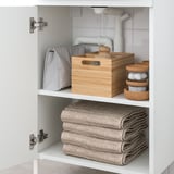 Un armario de baño contén toallas dobradas, unha caixa de madeira e organizadores de cortiza redonda, proporcionando almacenamento organizado.