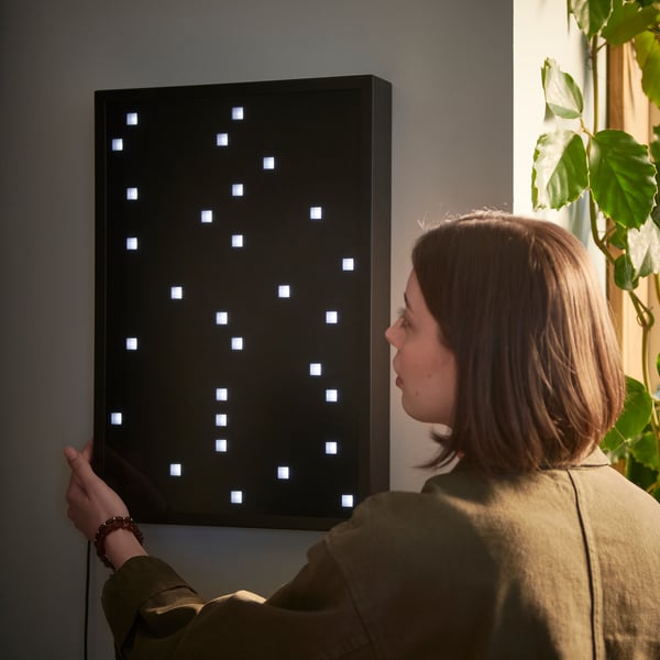 Unha muller interactúa cunha lámpada de parede LED negra e rectangular OBEGRÄNSAD cunha cuadrícula de cadrados iluminados.