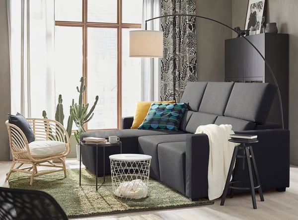 Sala de estar moderna con sofá seccional gris ODENSLUNDA, almofadas de cores, cadeira de vimbio e gran xanela con cortinas con patróns.