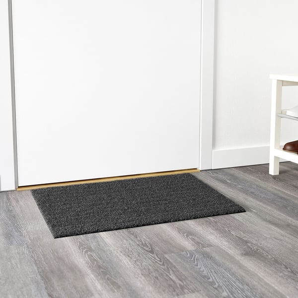 Alfombra rectangular gris escuro colocada contra unha parede branca nun chan de madeira, deseñada para uso exterior.