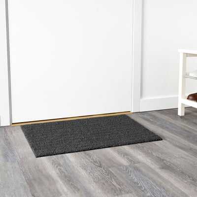Alfombra rectangular gris escuro colocada contra unha parede branca nun chan de madeira, deseñada para uso exterior.