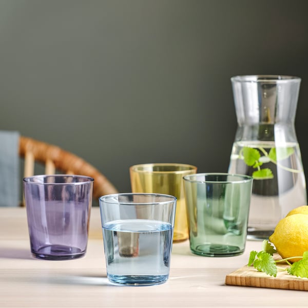 Catro vasos de cores e unha carafa con auga e franxas de limón sobre unha mesa.