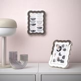 Estante branco con vasos decorativos de prata e dúas pezas de arte abstracta enmarcadas. Un marco apóiase no andel, o outro colga nunha parede rosa.
