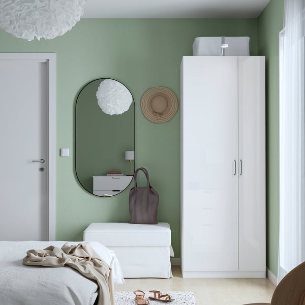 Dormitorio minimalista con armario branco PAX, espello e paredes verdes. Cama con liño branco e manta beige. A decoración inclúe lámpada, sombreiro e bolsa.