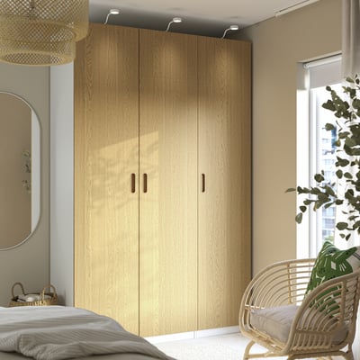 Dormitorio minimalista con gran armario PAX de carballo claro, cadeira de vimbio e plantas colgantes. Paredes beige suaves, espello grande e paralume de lámpada tecido.