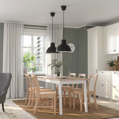 Comedor moderno con mesa branca, seis cadeiras, dúas lámpadas negras, gran xanela, zona de cociña acolledora.