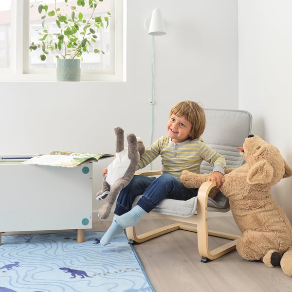 Neno sentado nunha mini cadeira de brazos POÄNG cun peluche, xunto a unha mesa con libros nunha habitación acolledora.